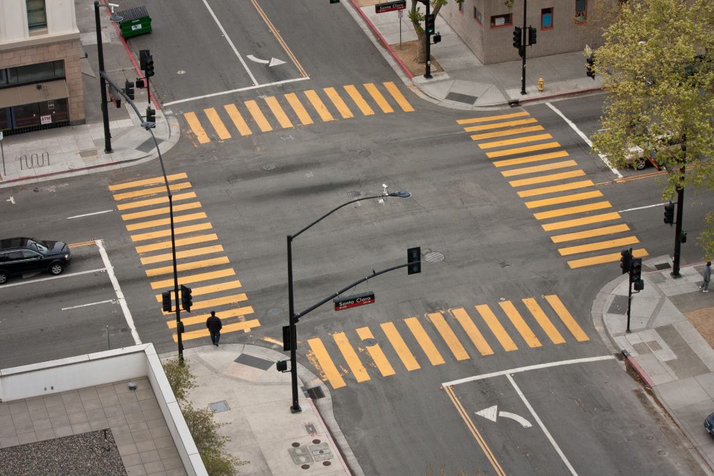 Aerial view of an urban intersection with bright yellow crosswalks. A lone pedestrian walks, while minimal traffic suggests a quiet city scene.